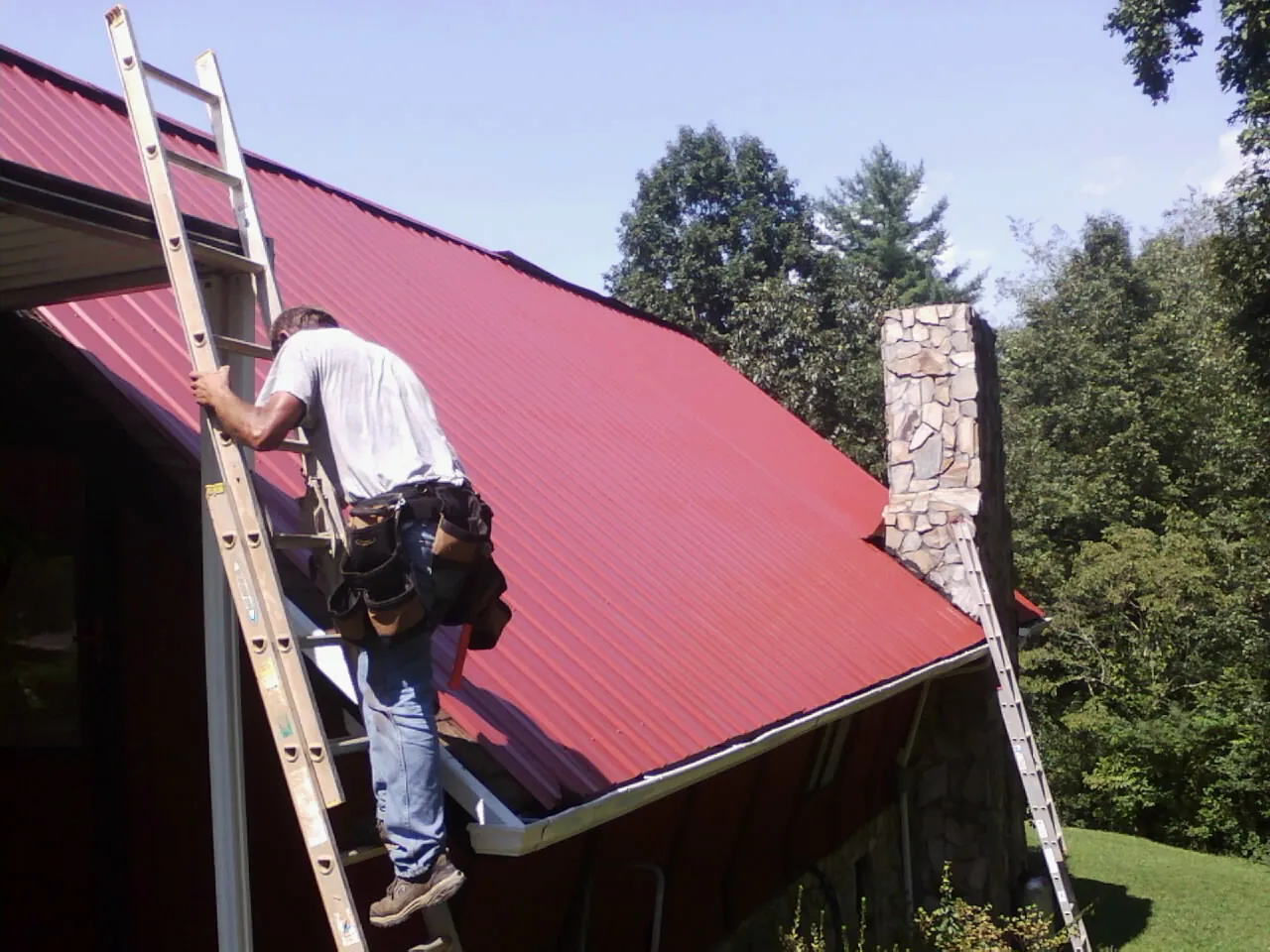 Roofer on a ladder beside a red metal roof and stone chimney — Soffit & Fascia Repair by Roofing Brothers in Metropolis